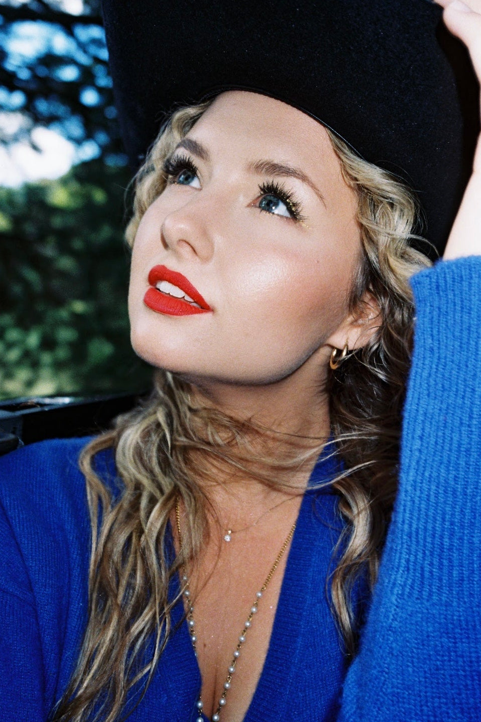 Woman wearing a black hat and blue sweater with red lipstick, sitting outdoors.